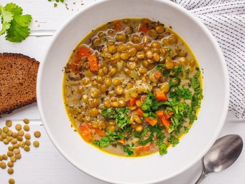 Aus der Vogelperspektive: Ein Teller Linsensuppe auf einem weißen Hintergrund. Drum herum liegen braune Linsen, eine Scheibe Brot und frische Petersilie