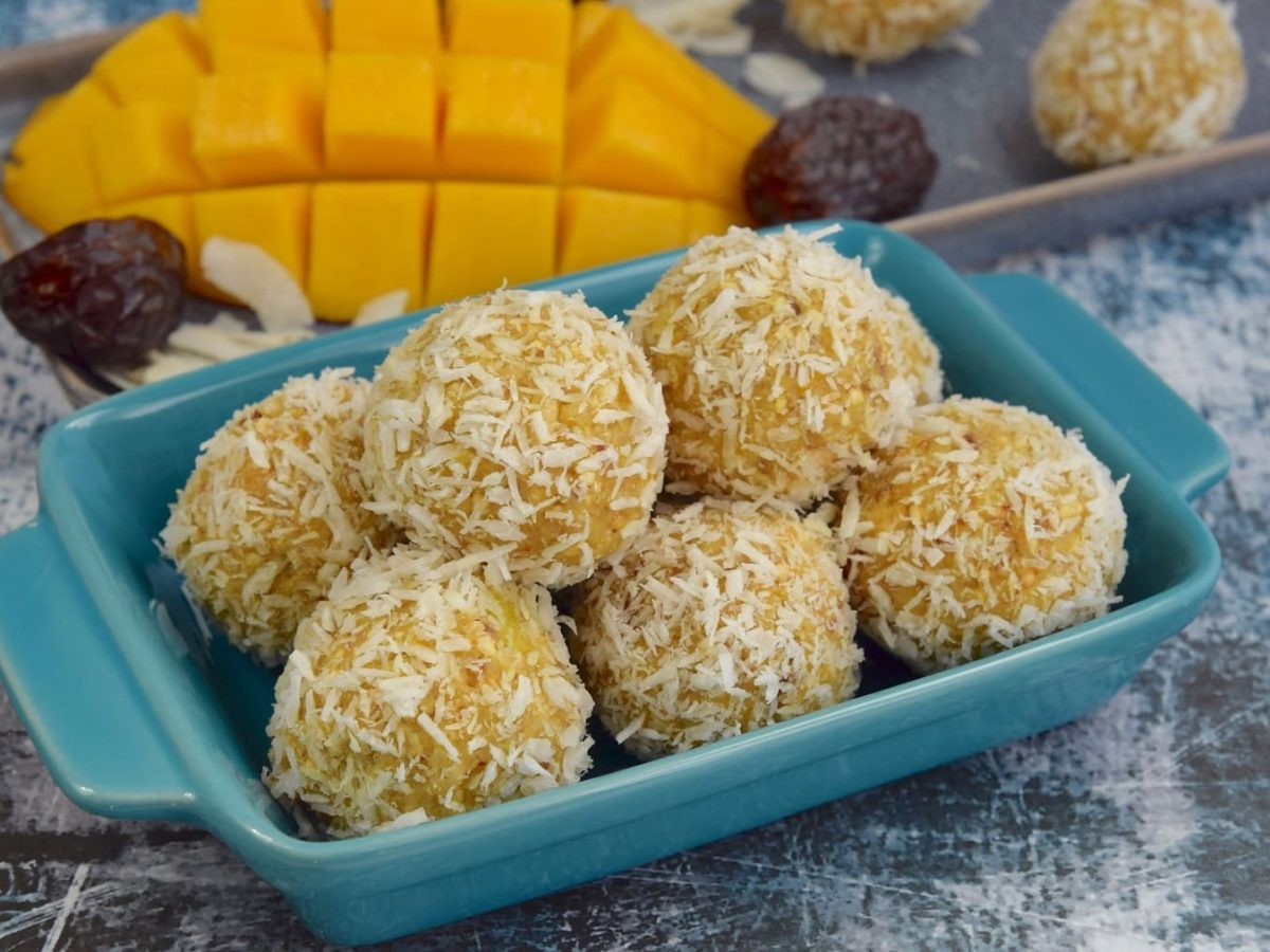 Ein paar Mango-Energy-Balls in einer blauen kleinen Auflaufform, dahinter eine angeschnittene Mango sowie Datteln.