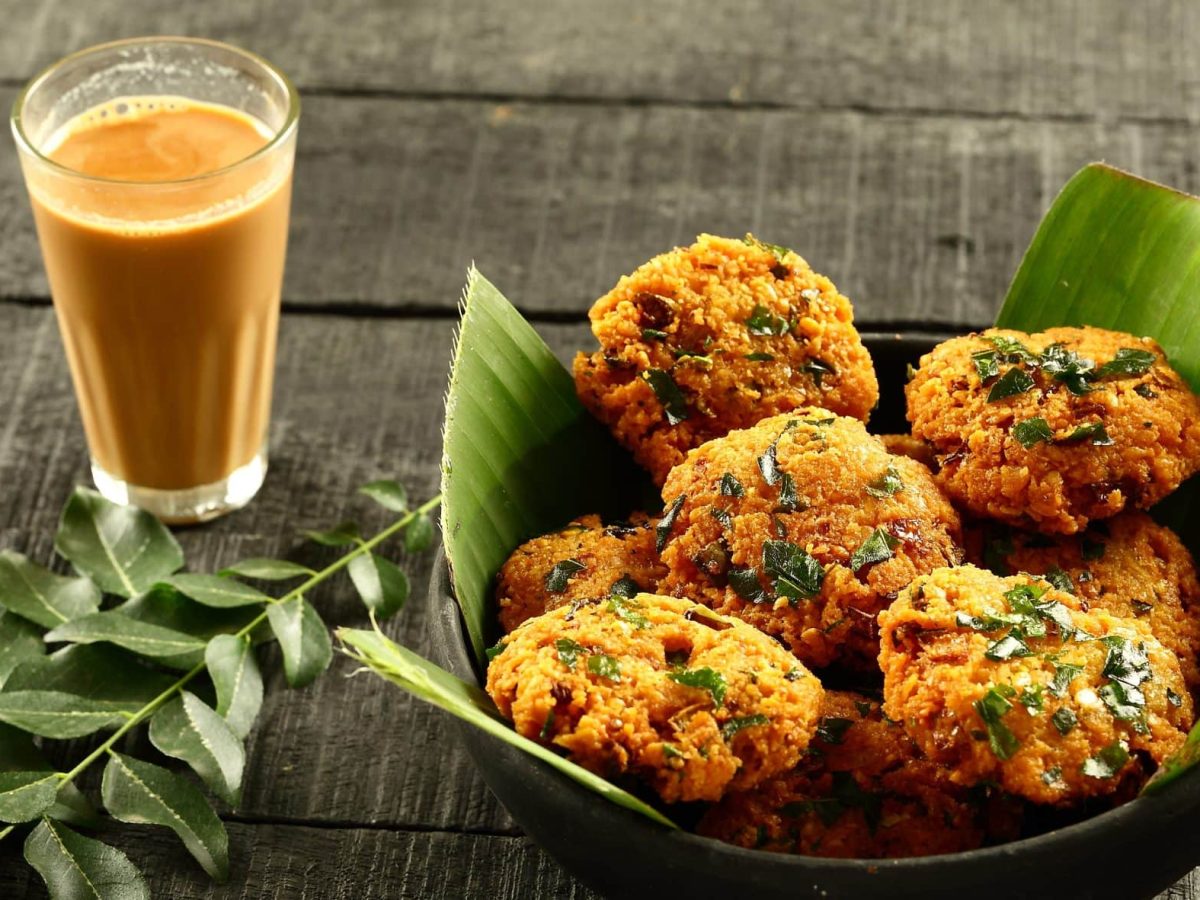 Masala Dal Vada in schwarzer Schale mit Blättern auf grauem Tisch. Links Curryblätter als Deko. Ein Glas mit Masala Chai oben im Bild. Draufsicht.