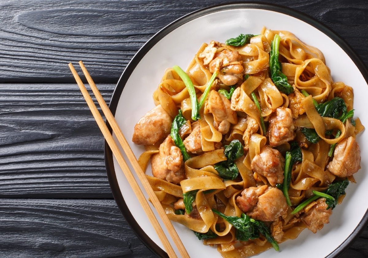 Gebratene Reisbandnudeln mit Hähnchen und Pak-Choi gebraten. Der Untergrund ist ein dunkler Holztisch.