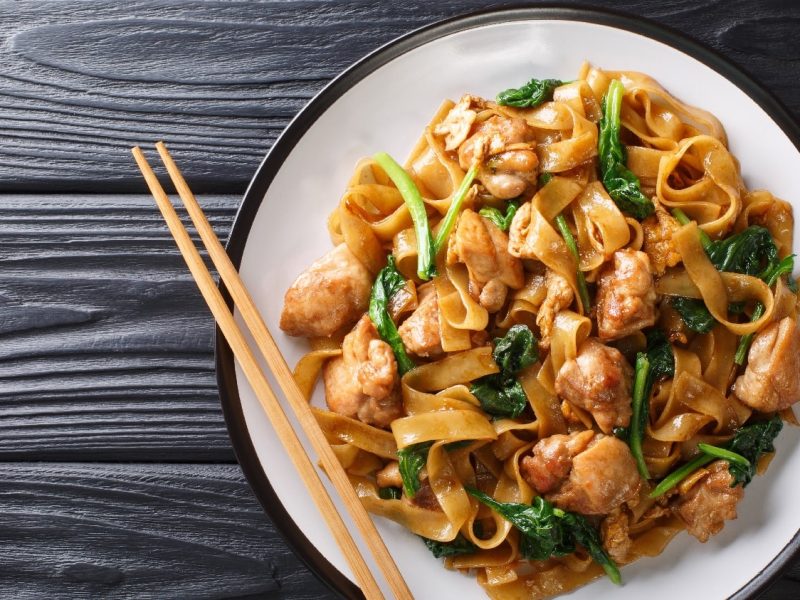 Gebratene Reisbandnudeln mit Hähnchen und Pak-Choi gebraten. Der Untergrund ist ein dunkler Holztisch.