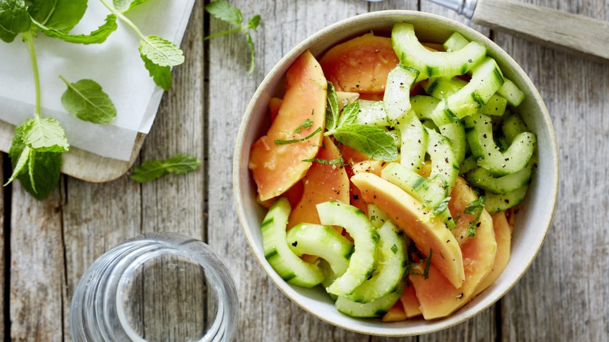 Papaya-Gurken-Duo in einer Schüssel neben etwas Minze vor Holzhintergrund.