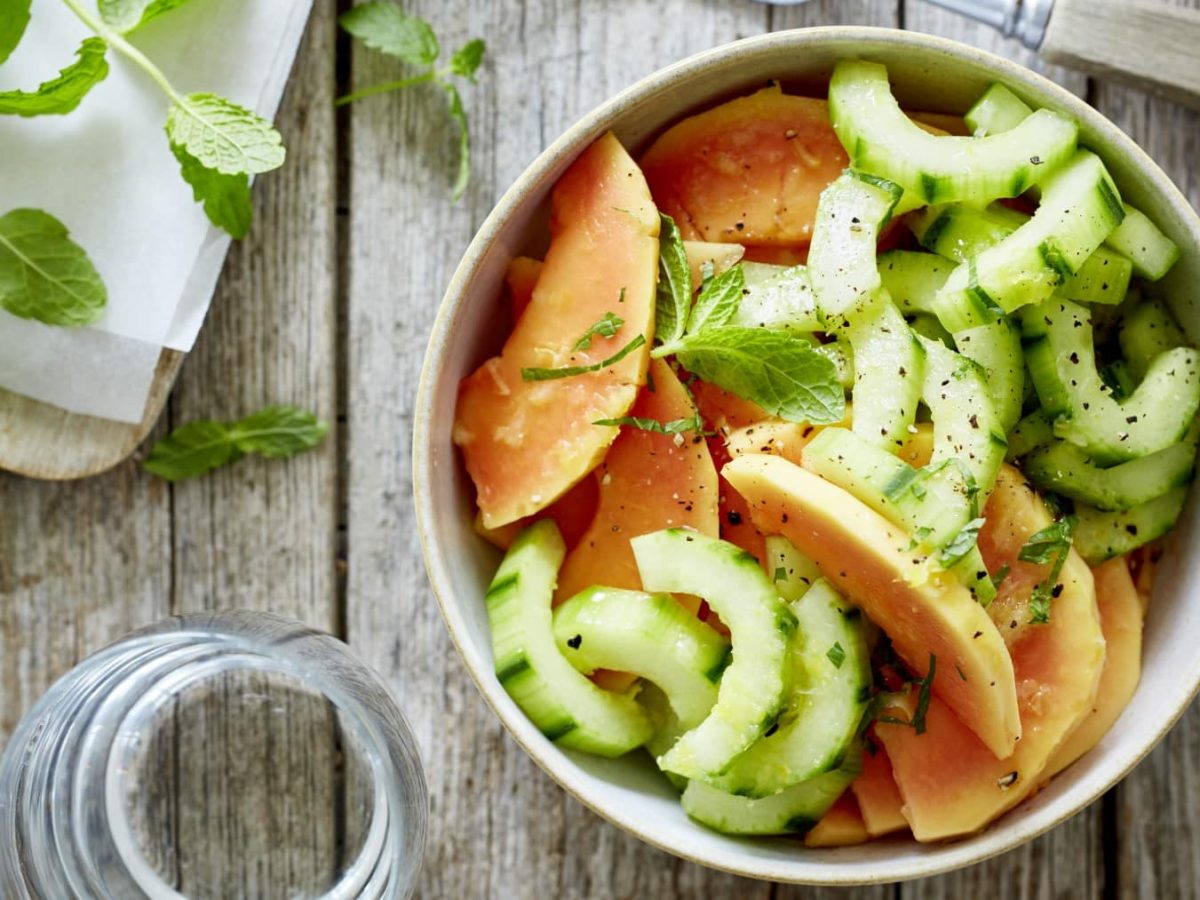 Papaya-Gurken-Duo in einer Schüssel neben etwas Minze vor Holzhintergrund.