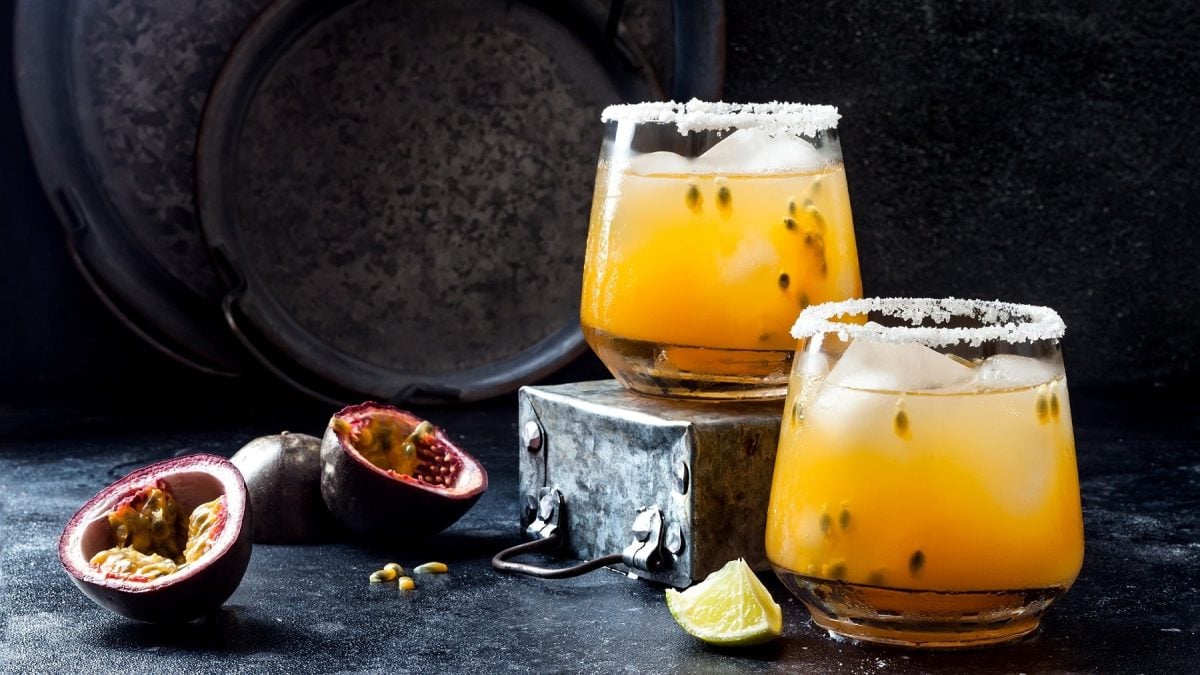 Zwei Gläser Passionfruit Paloma, ein gelber Cocktail mit Maracuja-Fruchtfleisch und Eis, daneben lieben halbierte Maracujas und Limettenspalten.