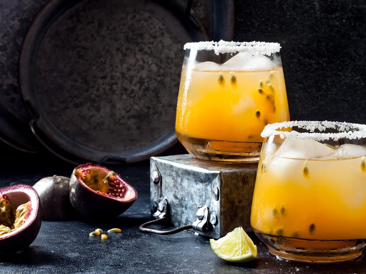 Zwei Gläser Passionfruit Paloma, ein gelber Cocktail mit Maracuja-Fruchtfleisch und Eis, daneben lieben halbierte Maracujas und Limettenspalten.