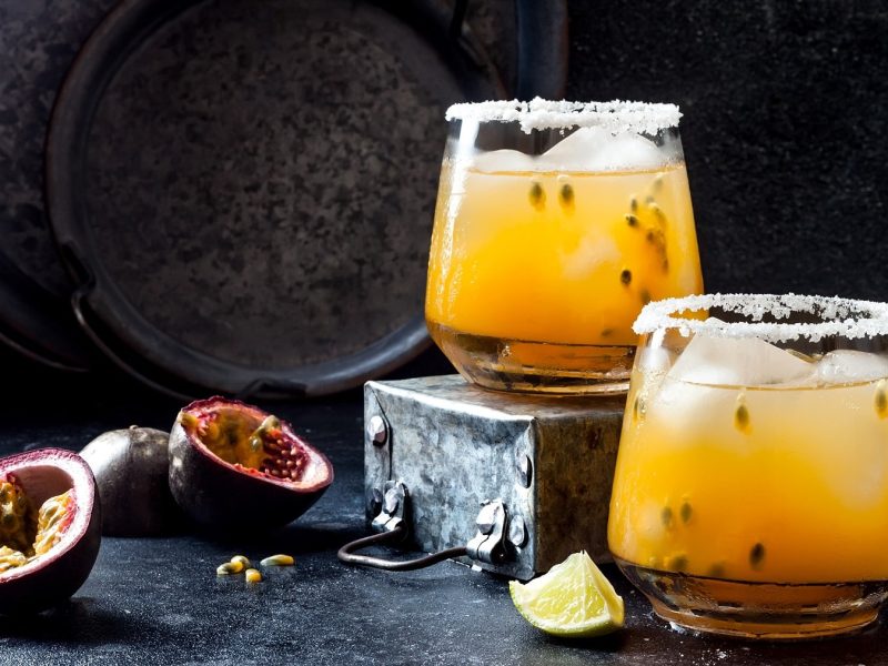 Zwei Gläser Passionfruit Paloma, ein gelber Cocktail mit Maracuja-Fruchtfleisch und Eis, daneben lieben halbierte Maracujas und Limettenspalten.