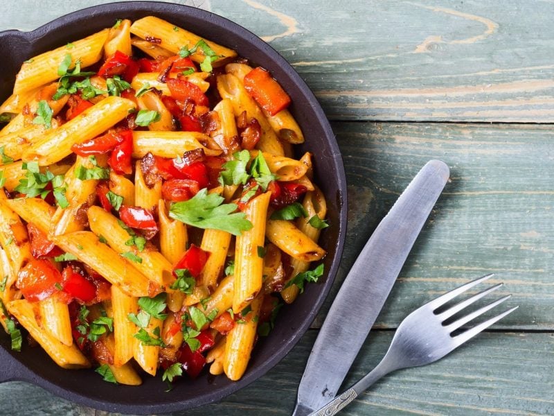 Eine gusseiserne Pfanne mit Penne mit gerösteter Knoblauch-Paprika-Sauce und Petersilie in der Draufsicht, daneben Messer und Gabel.