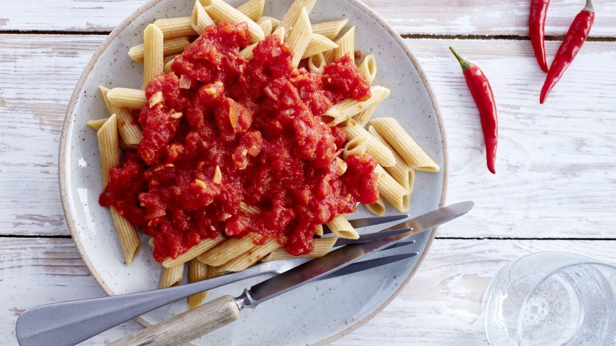 Chilisauce und Penne auf einem weißen Teller neben Besteck und Chilischoten vor hellem Hintergrund.