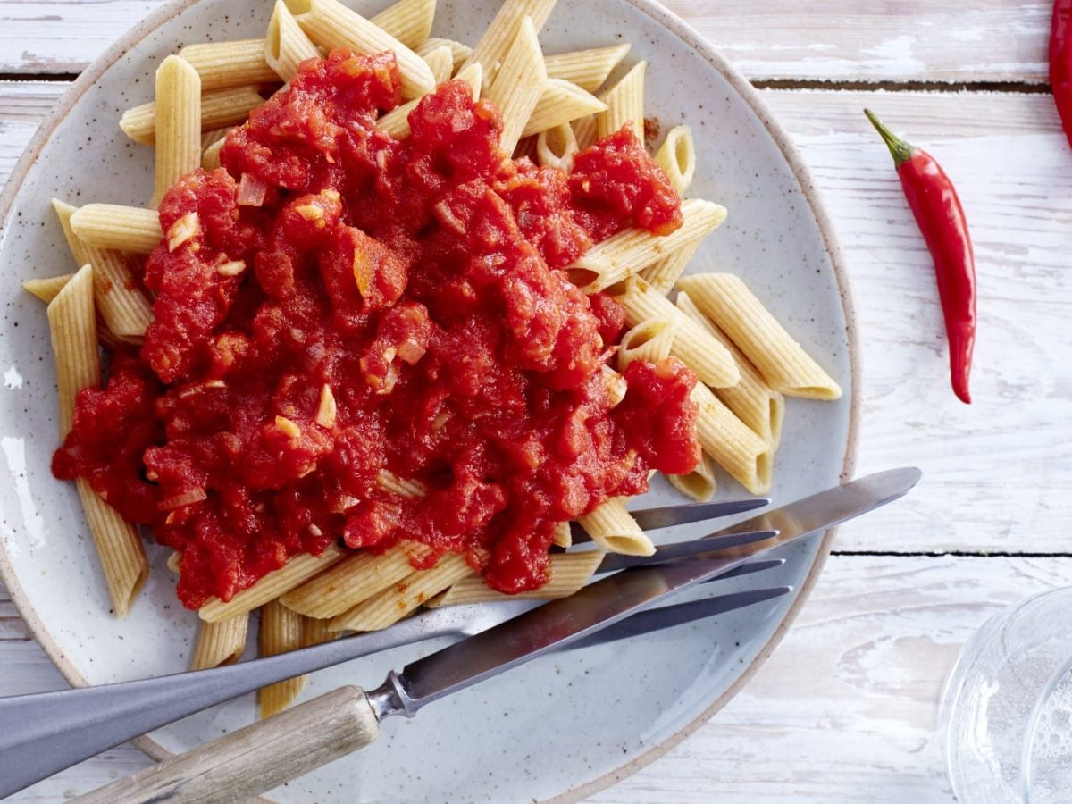 Chilisauce und Penne auf einem weißen Teller neben Besteck und Chilischoten vor hellem Hintergrund.