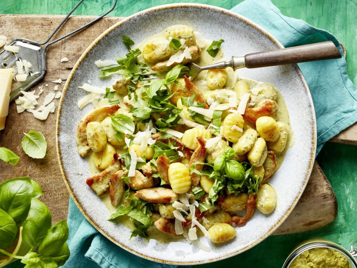 Pesto-Hähnchen-Geschnetzeltes mit Gnocchi auf einem Teller neben Parmesan und Basilikum auf einem Holzbrett vor grünem Hintergrund.