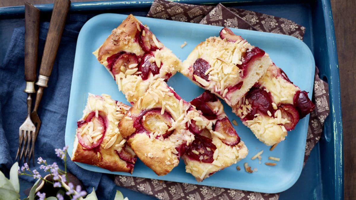 Einige Stücke Pflaumen-Butterkuchen auf einem hellblauen Teller neben Gabeln vor dunkelblauem Hintergrund.