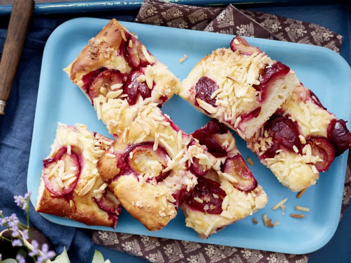 Einige Stücke Pflaumen-Butterkuchen auf einem hellblauen Teller neben Gabeln vor dunkelblauem Hintergrund.