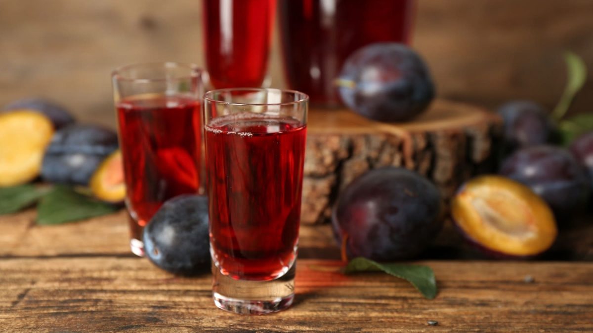 Shotgläser mit rotem Pflaumenlikör neben einigen Pflaumen vor Holzhintergrund.