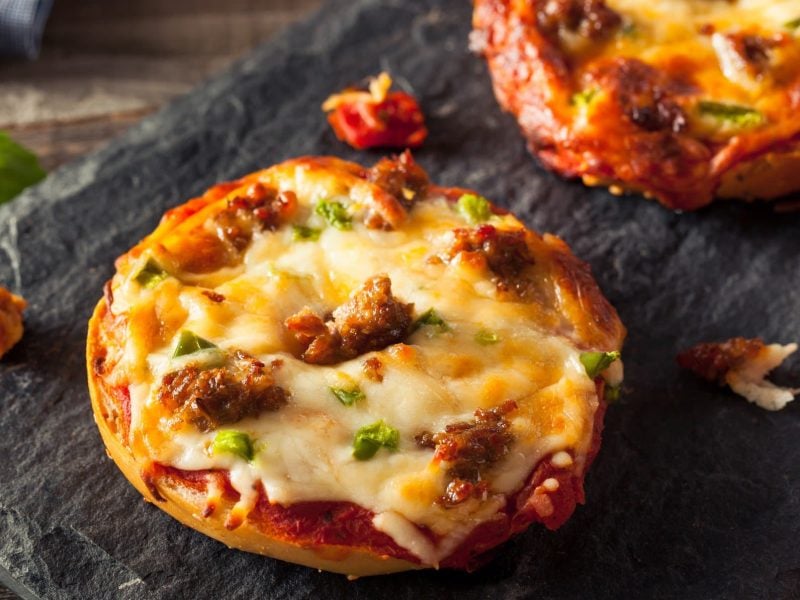 Eine schwarze Schieferplatte, mit zwei Pizza Bagels auf einem Holzuntergrund. Daneben ein Zweig frischer Basilikum.