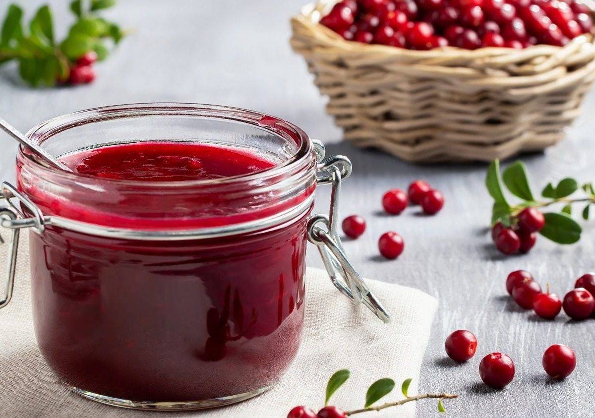 Frontalsicht: EIn Glas Preiselbeer-Konfitüre steht auf einem Tuch. drum herum liegen Preiselbeeren. Im Hintergrund steht ein Bastkorb mit frischen Preiselbeeren.