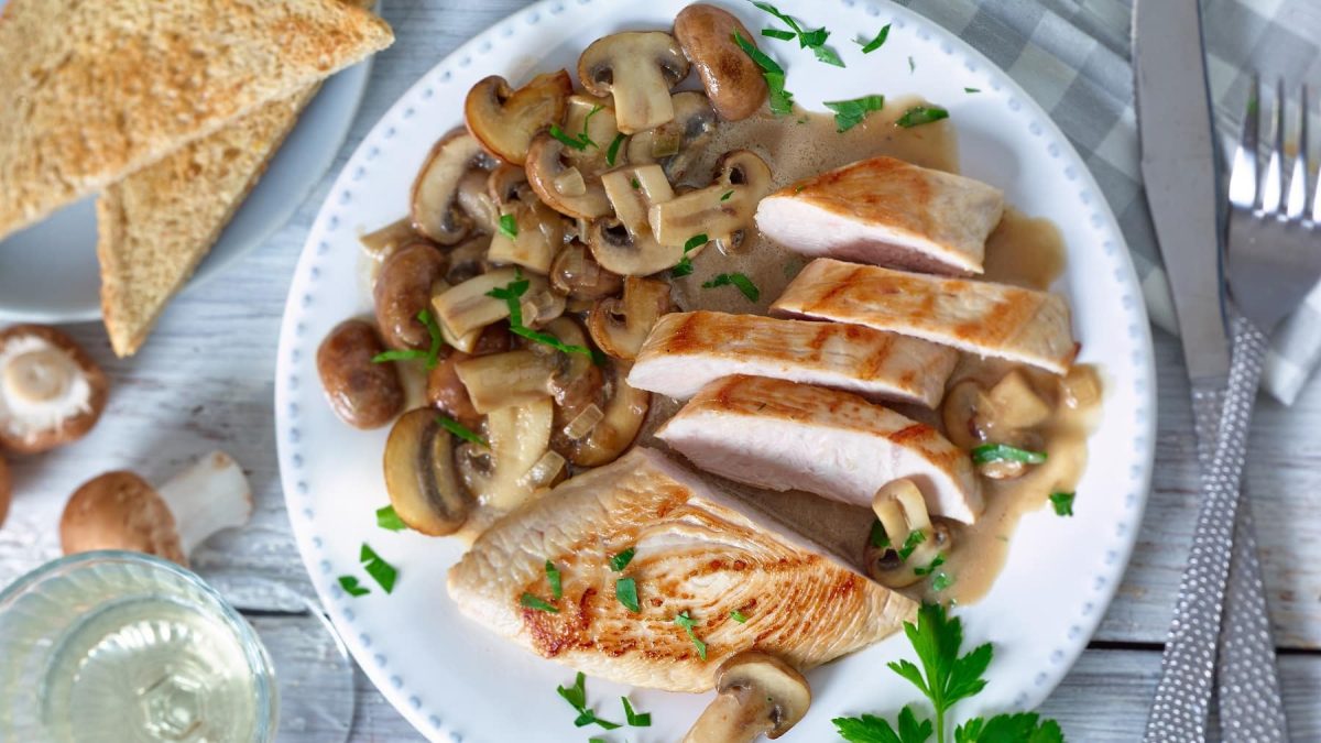 Putenschnitzel mit Champignons auf weißem Teller garniert mit Kräutern neben Brot und Champignons vor hellem Hintergrund.