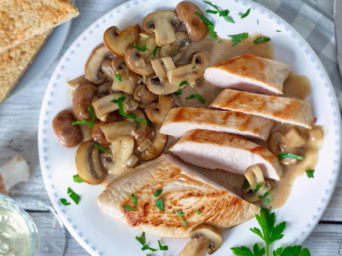 Putenschnitzel mit Champignons auf weißem Teller garniert mit Kräutern neben Brot und Champignons vor hellem Hintergrund.