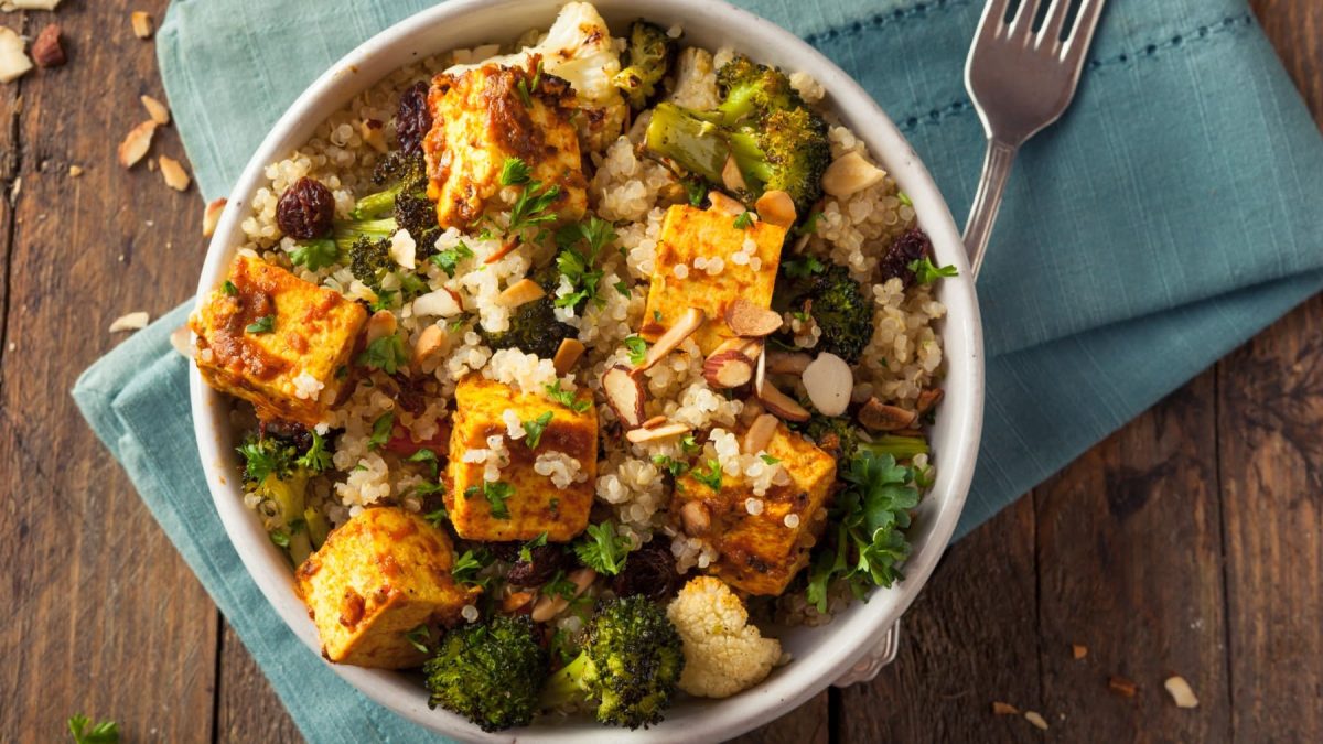 Quinoa-Tofu-Bowl in einer Schüssel und eine Gabel auf blauem Stoff vor Holzhintergrund.