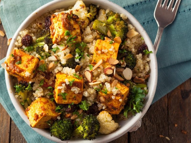 Quinoa-Tofu-Bowl in einer Schüssel und eine Gabel auf blauem Stoff vor Holzhintergrund.