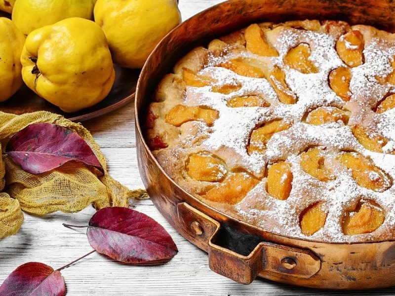 Aus der Vogelperspektive: Ein runder Quittenkuchen steht auf einem hellen Untergrund. Drum herum liegen Blätter von Bäumen sowie frische Quitten.