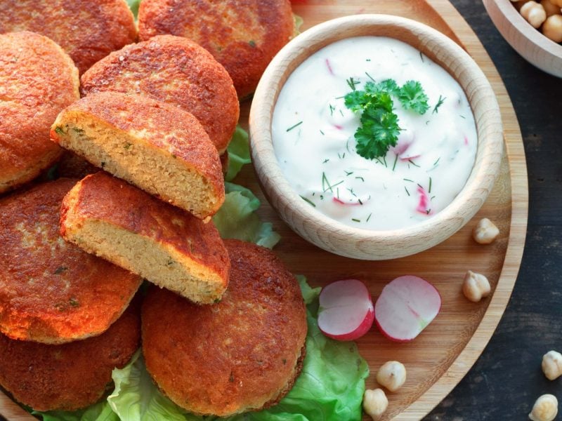 Radieschen-Raita mit Kichererbsenküchlein und Blattsalat auf Holzteller. Daneben Kichererbsen und Radieschen. Draufsicht.