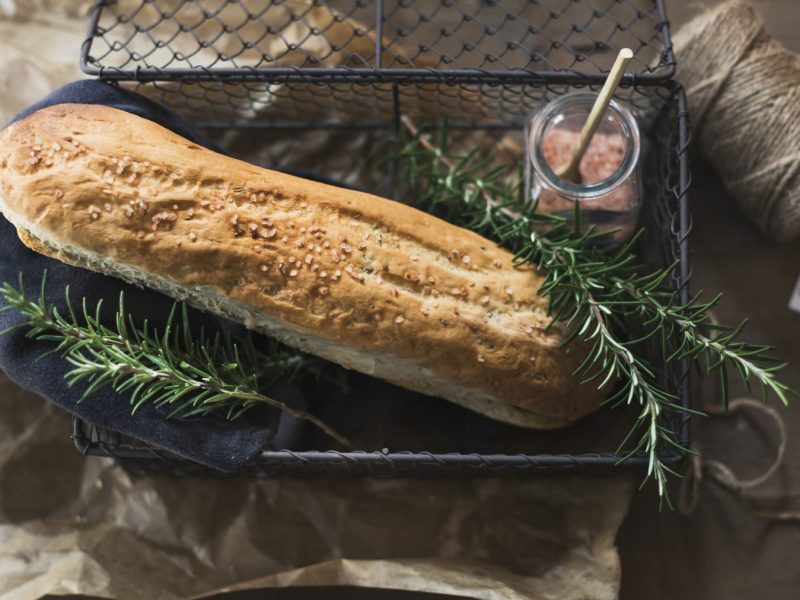 Rosmarin-Baguette dekoriert mit Rosmarinzweigen und rosa Steinsalz in einem schwarzen Drahtkorb vor braunem Hintergrund.