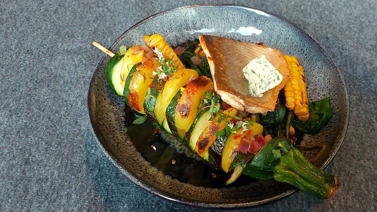 Eine blauer tiefer Teller, mit dem Spieß und dem Fisch auf Spinat. Der Untergrund ist dunkelgrau.