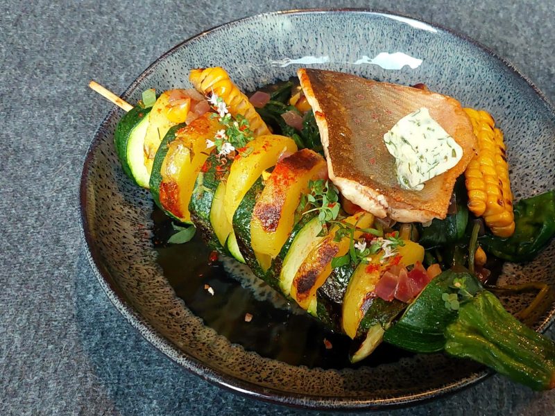 Eine blauer tiefer Teller, mit dem Spieß und dem Fisch auf Spinat. Der Untergrund ist dunkelgrau.