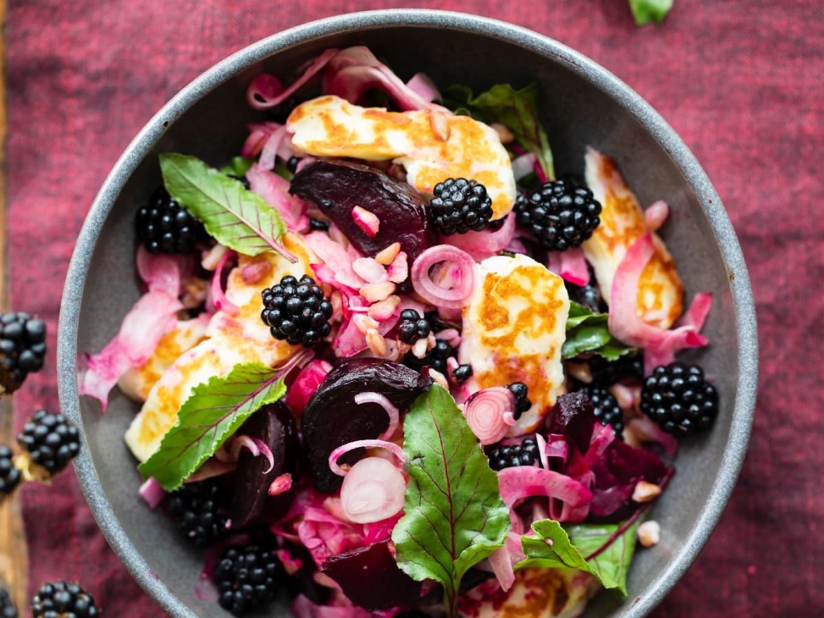 Eine graue Schale mit dem Halloumi-Salat auf einer weinroten Tischdecke. Unten links ist ein Stück vom alten Holztisch und ein Brombeerstrauch zu sehen.