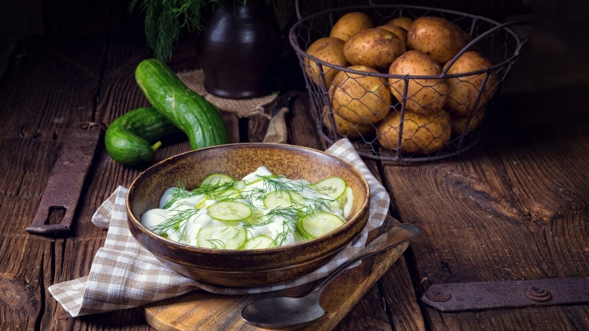 Schräge Ansicht von oben: Ein Salat mit Kartoffeln und Gurken steht auf einem Tisch. Dahinter liegen Kartoffeln und Salatgurken.