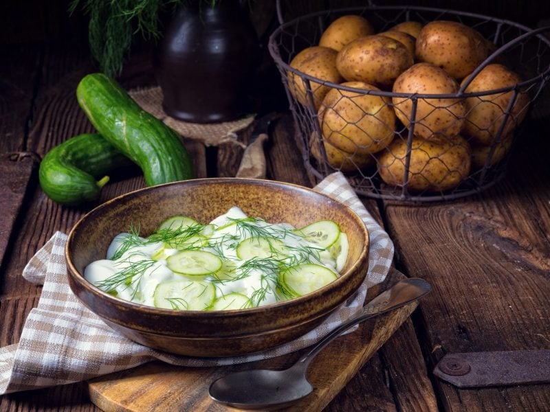 Schräge Ansicht von oben: Ein Salat mit Kartoffeln und Gurken steht auf einem Tisch. Dahinter liegen Kartoffeln und Salatgurken.