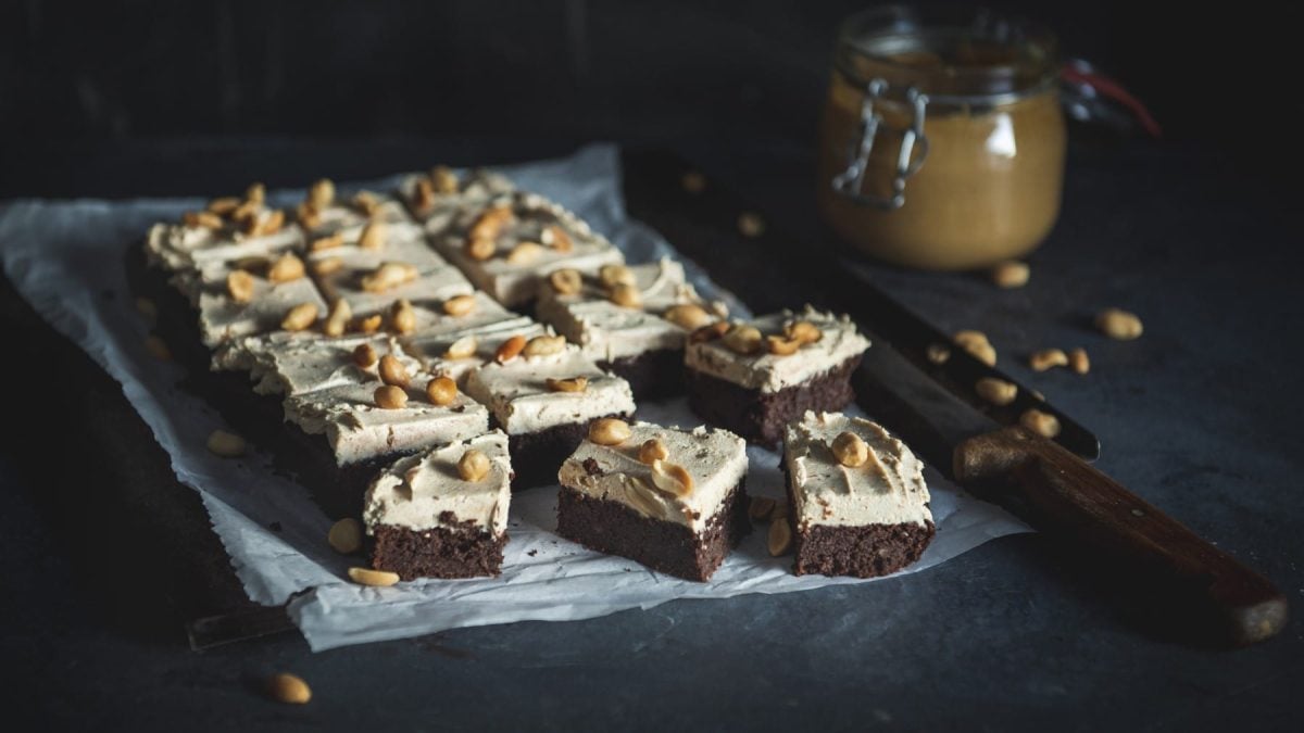 Schoko-Brownies mit Peanut-Butter-Frosting garniert mit gerösteten Erdnüssen auf Backpulver mit einem Glas Erdnussbutter im dunklen Hintergrund.