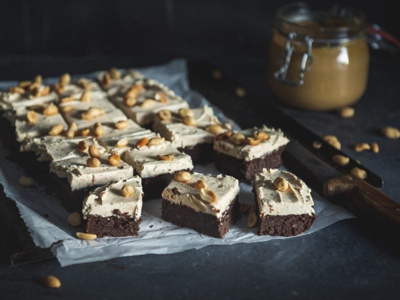 Schoko-Brownies mit Peanut-Butter-Frosting garniert mit gerösteten Erdnüssen auf Backpulver mit einem Glas Erdnussbutter im dunklen Hintergrund.