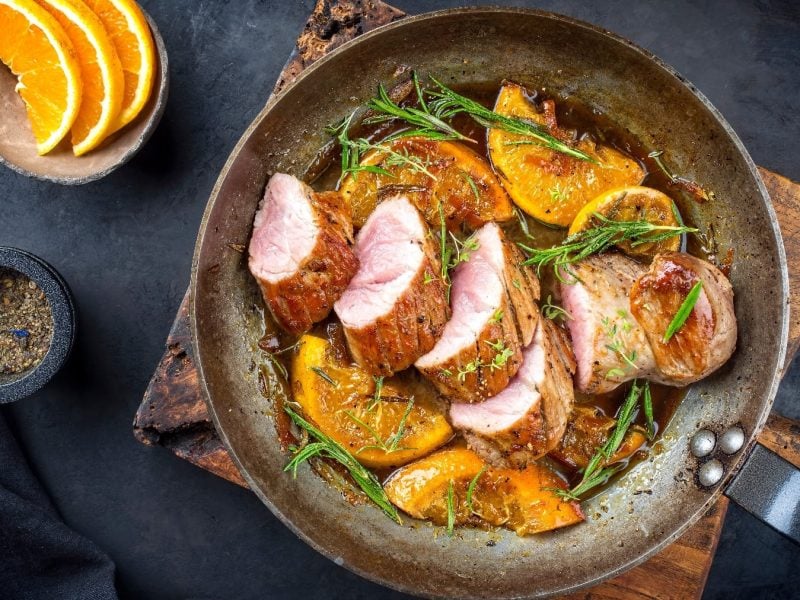 Eine alte Pfanne mit dem geschnittenen Schweinefilet auf der Orangen-Senfsauce und grünen Kräutern. Die Pfanne steht auf einem Holzschneidebrett und dieses wiederum auf einem dunkelblauen Tisch mit einer Schale von Orangen.