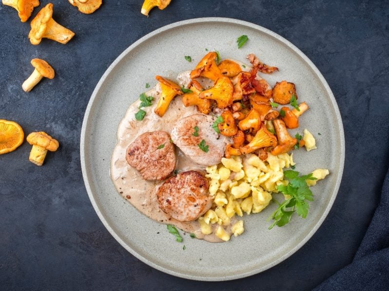 Ein grauer Teller mit den Schweinemedallions an Rahmsauce, Pfifferlingen und Knöpfle. Alles auf einem dunkelblauem Untergrund mit Pfifferlingen in der Ecke oben links.