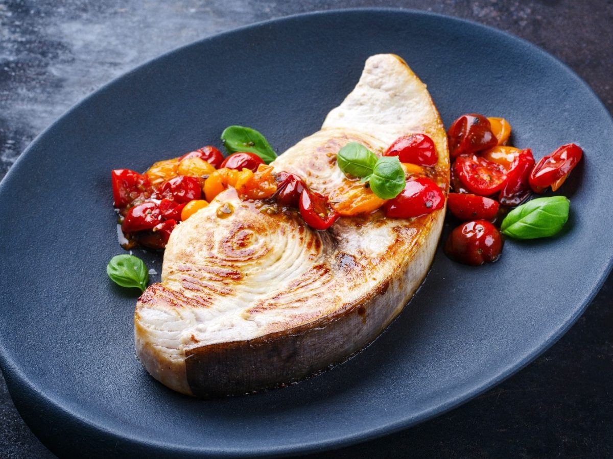 Ein schwarzer Teller mit dem Schwertfisch und Tomatenragout auf einem dunklen Untergrund.