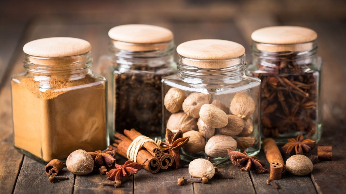 Vier Gläser mit Zutaten für ein selbst gemachtes Kaffeegewürz, darunter Nelken und Muskatnüsse.