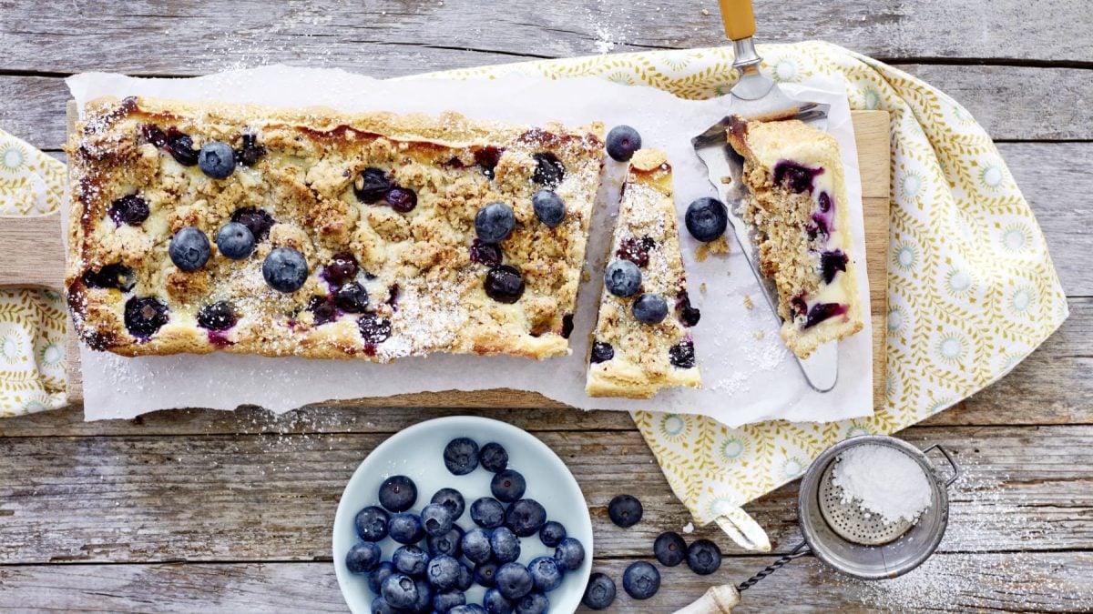 Shortbread-Heidelbeer-Tarte liegt auf einem länglichen Brett. Daneben steht eine Schale mit Heidelbeeren und ein Sieb mit Puderzucker.