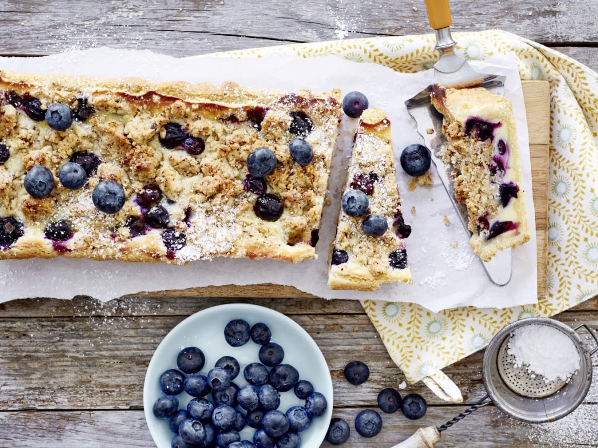 Shortbread-Heidelbeer-Tarte liegt auf einem länglichen Brett. Daneben steht eine Schale mit Heidelbeeren und ein Sieb mit Puderzucker.