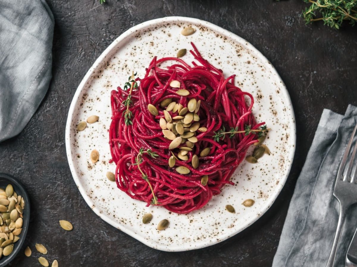 Spaghetti mit Rote-Bete-Pesto auf einem braunweiß gemusterten Teller. Daneben liegen verschiedene Zutaten sowie Besteck und Küchentücher.