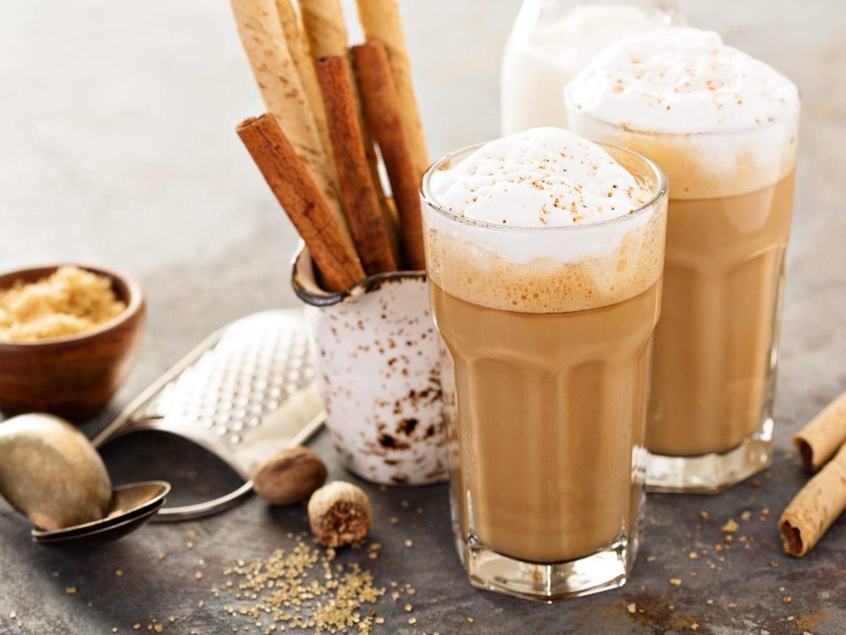 Zwei Gläser Spiced Latte mit Milchschaum, drumherum Gewürze wie Zimtstangen und Muskatnüsse.