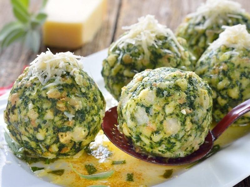 5 Spinatknödel liegen auf einer weißen Platte. IM Hintergrund ist ein Stück Käse-