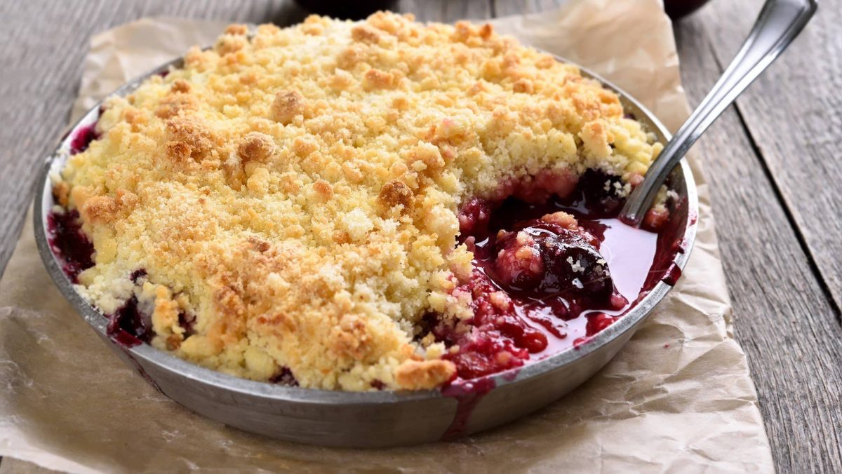 Steinobstcrumble in Auflaufform mit Meltalllöffel auf Backpapier und Holztisch. Draufsicht.