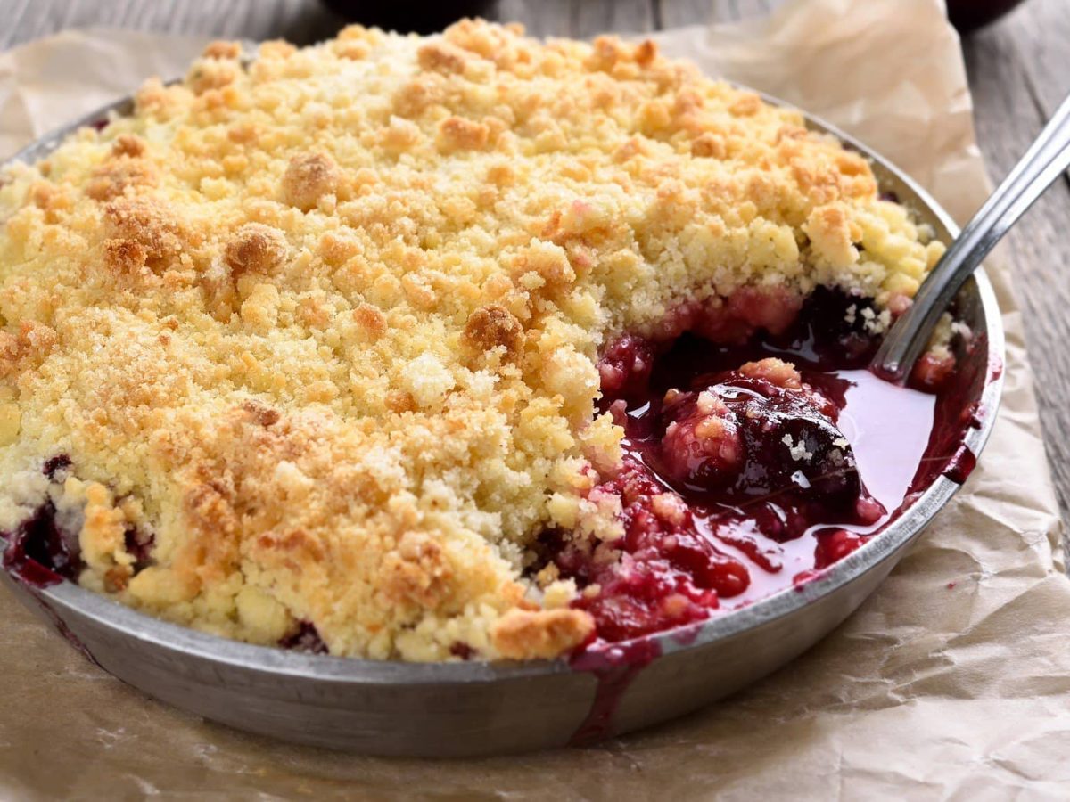 Steinobstcrumble in Auflaufform mit Meltalllöffel auf Backpapier und Holztisch. Draufsicht.