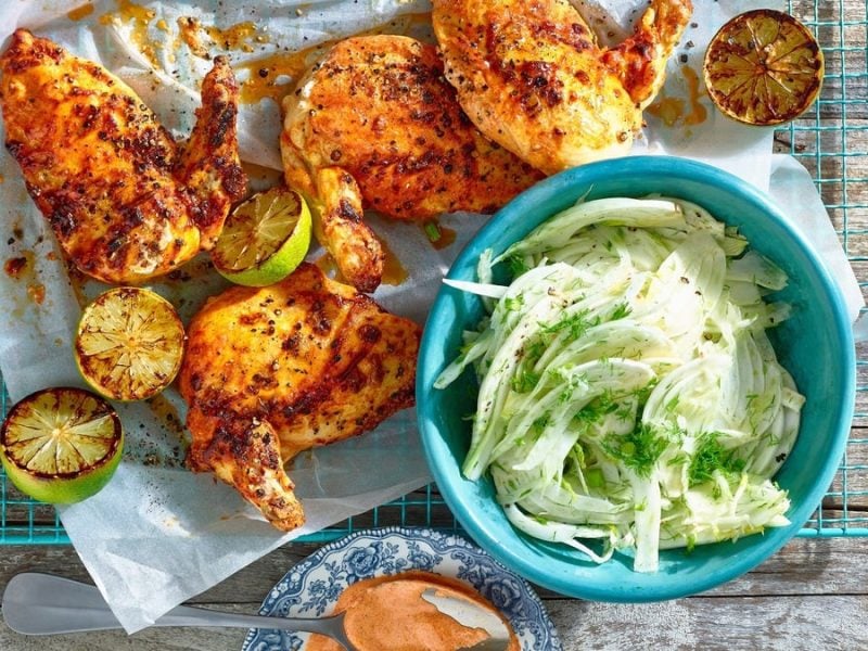 Mehrere Stücke Tandoori-Hähnchen mit Fenchel daneben in einer Schüssel auf einem blauen Gitter vor Holzhintergrund.