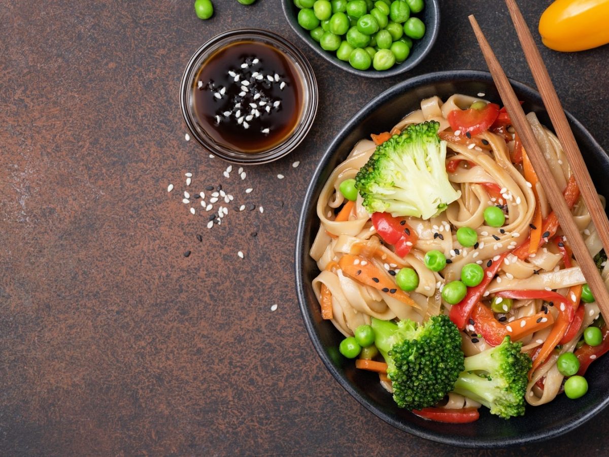 Ein dunkler Teller Teriyaki-Pasta mit Brokkoli, Paprika, Möhren und Erbsen, darauf Essstäbchen, daneben Gemüse, alles Draufsicht.