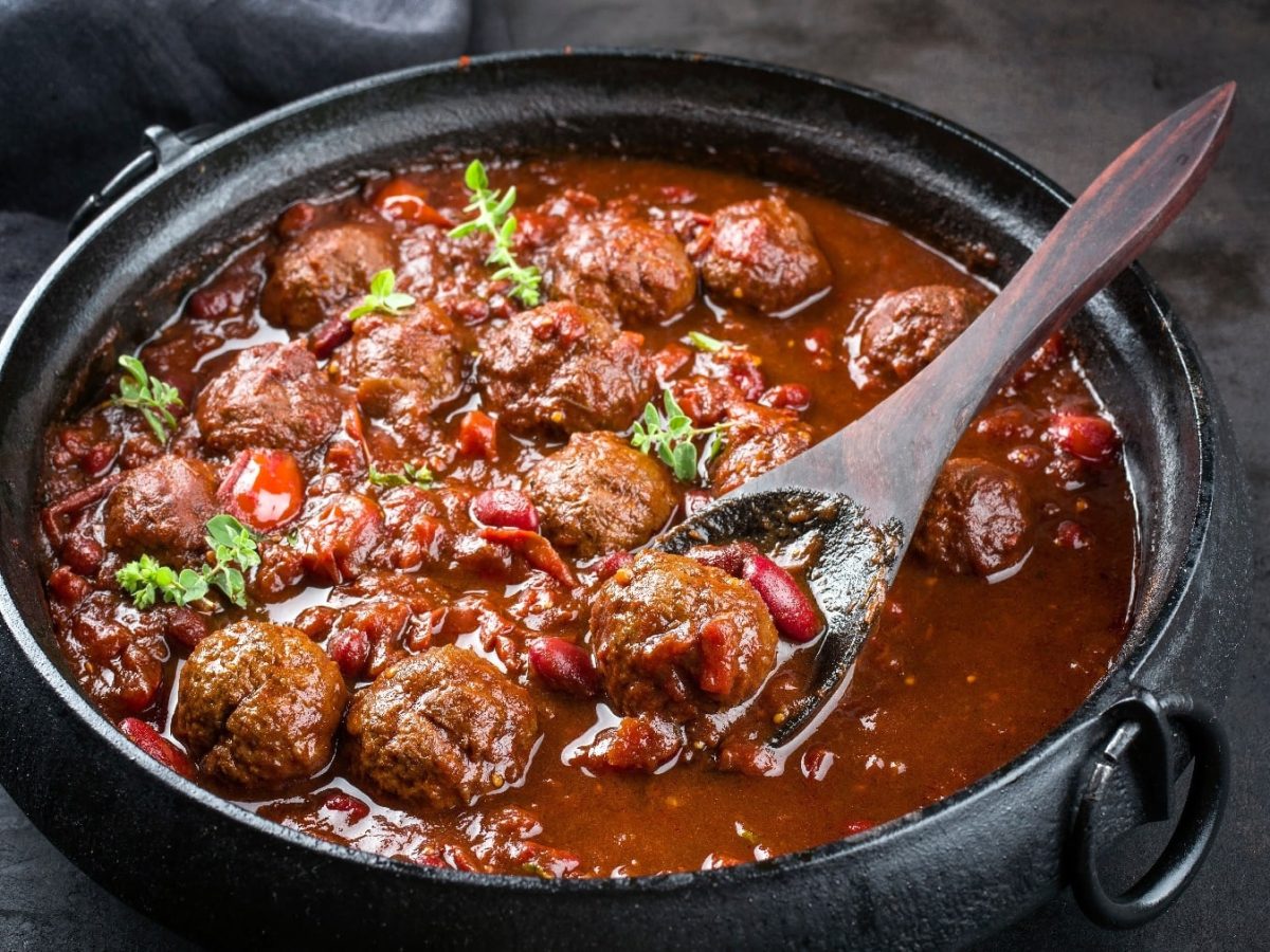 Ein schwarzer Topf mit den Hackbällchen in einer Tomatensauce mit Bohnen und PAprika auf schwarzem Untergrund.