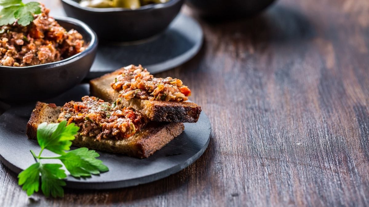 Zwei Brotscheiben mit Tomaten-Tapenade auf einem Teller, daneben Petersilie und eine Schale mit der Tapenade.