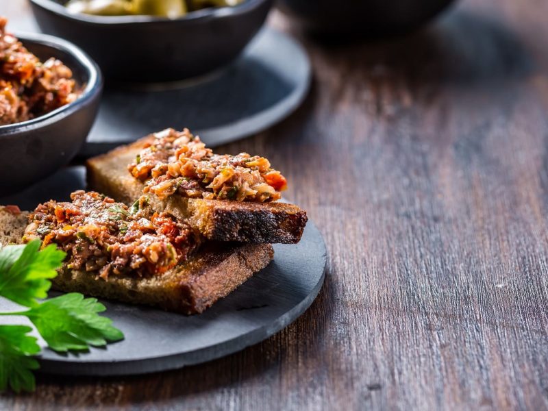 Zwei Brotscheiben mit Tomaten-Tapenade auf einem Teller, daneben Petersilie und eine Schale mit der Tapenade.