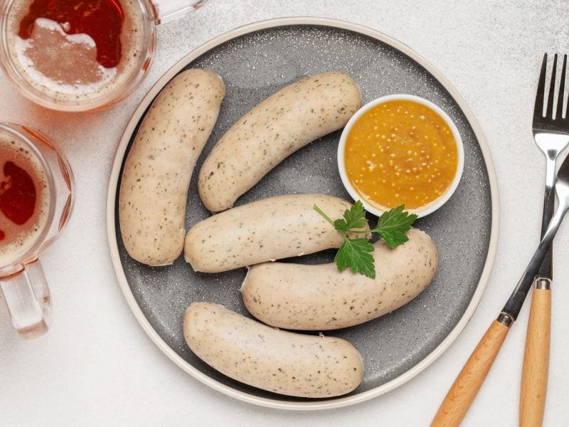 Fünf vegane Weißwürste mit einer Schale süßem Senf auf einem Teller, daneben zwei Gabeln und zwei Biergläser. Draufsicht.